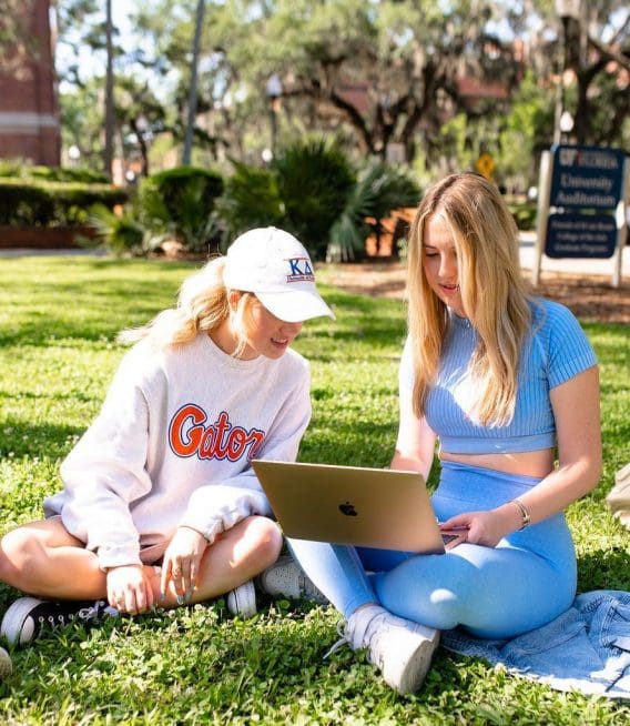 Two young women sit on the grass off campus, looking at a laptop together. One wears a Gators sweatshirt and white cap, the other a blue crop top and leggings. Campus buildings and trees can be seen in the background.