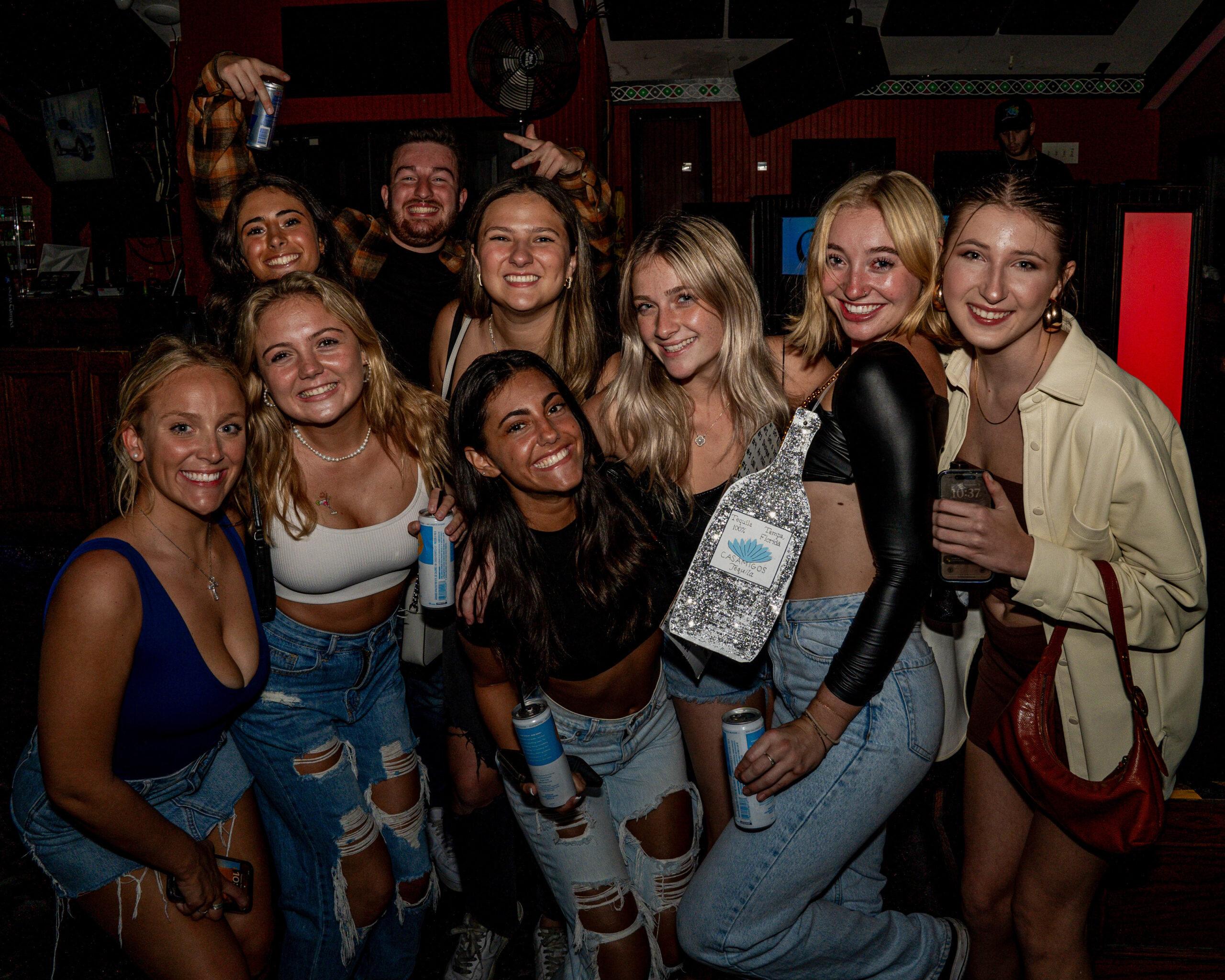 A group of smiling young adults pose together indoors at night, holding drinks and wearing casual outfits, mostly jeans and crop tops. The atmosphere suggests they are enjoying a night out at bars or a lively social gathering.